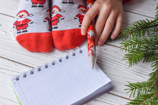 Een feestelijke kerst foto met vrouwelijke voeten in heldere rode sokken, een takje spar, een opmerking boek en een potlood op een witte houten vloer  - Foto, afbeelding