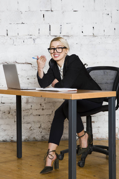 Jonge blonde schoonheid zakenvrouw zitten aan een bureau tafel met laptop, notitieboekje en glazen in pak. Bedrijfsconcept. - Foto, afbeelding