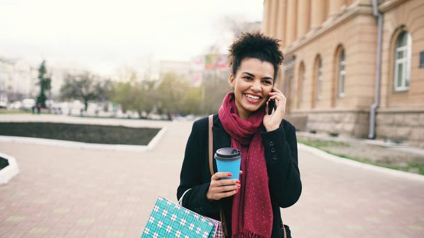 Attractive mixed race business woman talking smartphone and drinking coffee walks in city street with shopping bags - 写真・画像