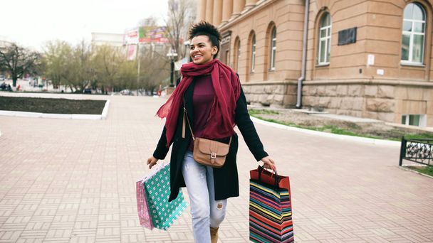 Attractive mixed race girl dancing and have fun while walking down the street with bags. Happy young woman walking after shopping on mall sale - Foto, Imagen