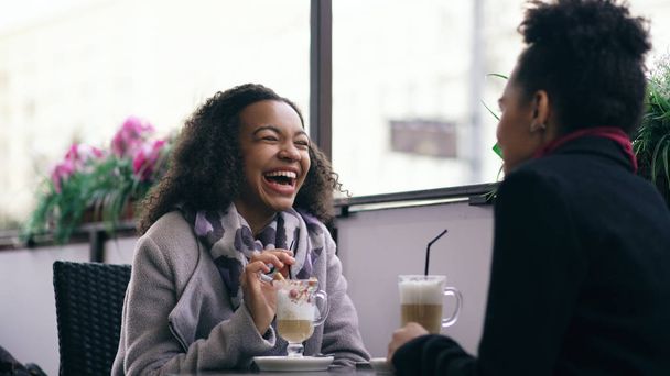 Two attractive mixed race women talking and drinking coffee in street cafe. Friends have fun after visiting mall sale - Foto, afbeelding