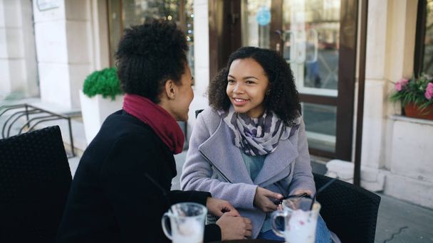 Two attractive mixed race women talking and drinking coffee in street cafe. Friends have fun after visiting mall sale - Foto, Bild