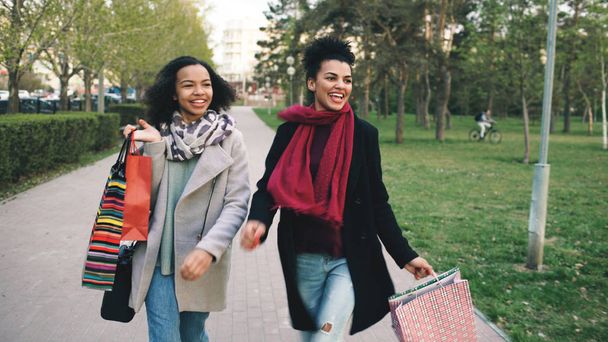 two attractive mixed race women dancing and have fun while walking down the park with shopping bags. Happy young friends walk after visiting mall sale - Foto, imagen