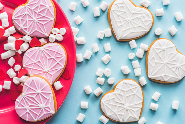 top view of glazed heart shaped cookies on pink plate with white marshmallow isolated on blue - Fotó, kép