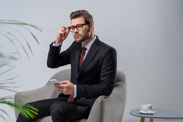 hombre de negocios sentado en sillón con teléfono inteligente y mirando hacia otro lado
 - Foto, Imagen