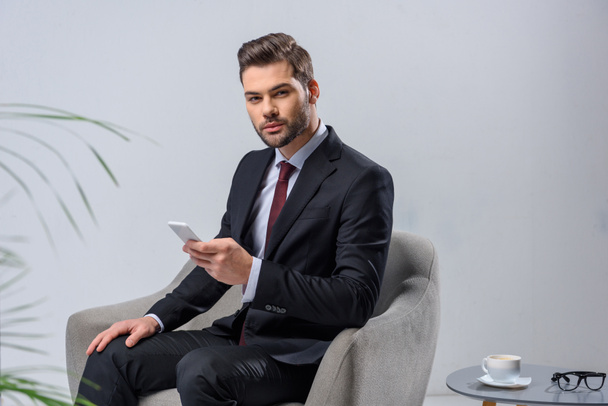 businessman sitting in armchair and holding smartphone - Zdjęcie, obraz