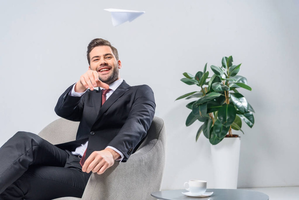smiling businessman sitting in armchair and throwing paper plane in office - Foto, afbeelding