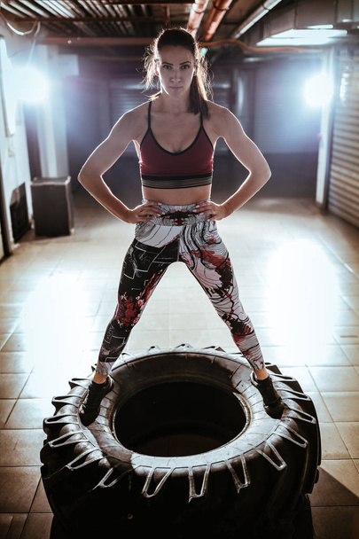 Young muscular woman standing on a tire and posing after cross fit training in the garage. Looking at camera. - Foto, immagini