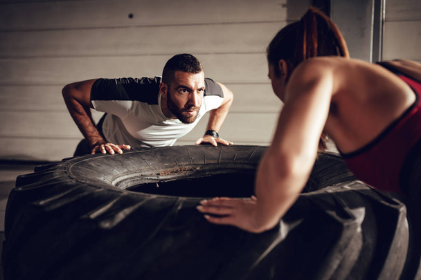 Young muscular couple doing push-up exercise on a tire at hard training in the garage. - Foto, imagen