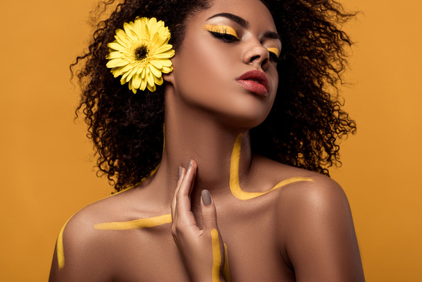 芸術的な化粧と髪でガーベラのスタイリッシュなアフリカ系アメリカ人女性に優しく触れる彼女の肌がオレンジ色の背景に分離 - 写真・画像