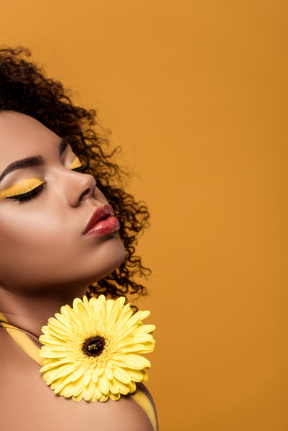 Young bright african american woman with artistic make-up holds yellow gerbera flower isolated on orange background - Photo, Image