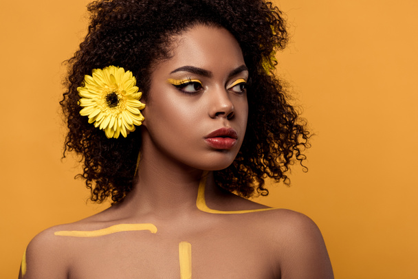 Joven mujer afroamericana sensual con maquillaje artístico y gerbera en el pelo mirando al lado aislado sobre fondo naranja
 - Foto, imagen