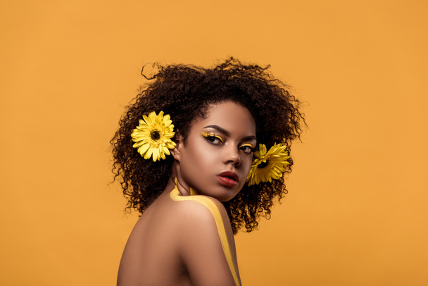 Jonge heldere african american vrouw met artistieke make-up en gerbera in haar geïsoleerd op een oranje achtergrond - Foto, afbeelding