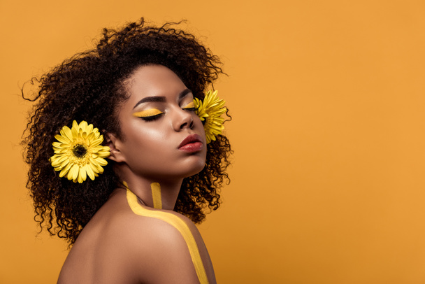 Jovem mulher americana africana brilhante com maquiagem artística e gerbera no cabelo sonhando isolado em fundo laranja
 - Foto, Imagem