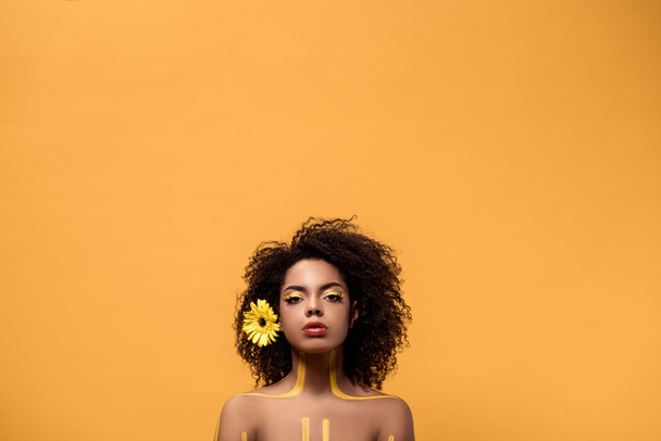 Nuori aistillinen afrikkalainen amerikkalainen nainen, jolla on taiteellinen meikki ja gerbera hiuksissa, katsoo kameraa, joka on eristetty oranssilla taustalla.
 - Valokuva, kuva