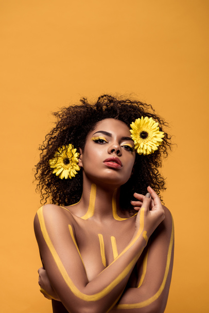 Jovem afro-americana com maquiagem artística e gerbera no cabelo de pé com braços cruzados isolados em fundo laranja
 - Foto, Imagem