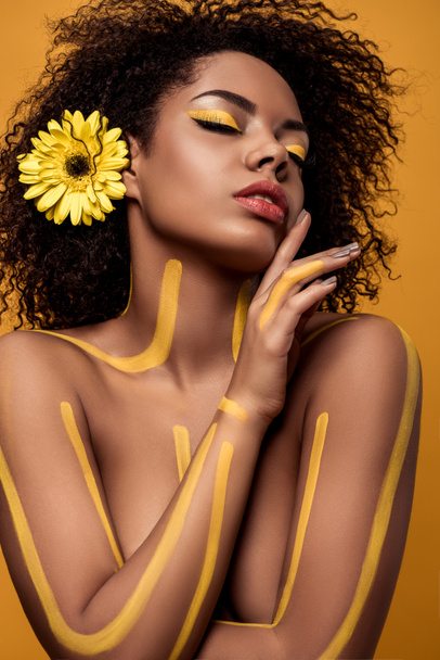 Joven mujer afroamericana sensual con maquillaje artístico y gerbera en pelo aislado sobre fondo naranja
 - Foto, imagen