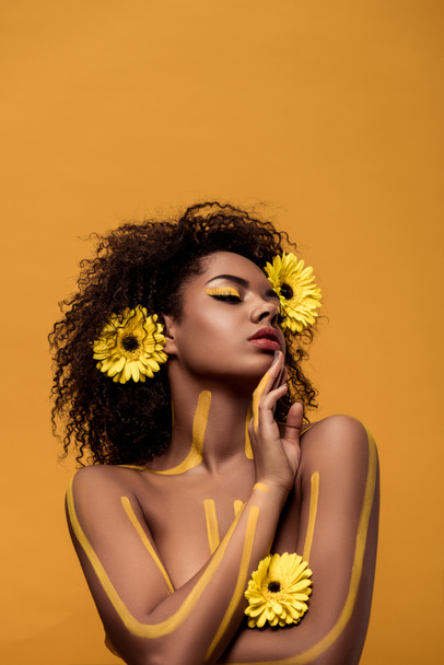 Mujer afroamericana tierna joven con maquillaje artístico y gerbera en pelo aislado sobre fondo naranja
 - Foto, imagen