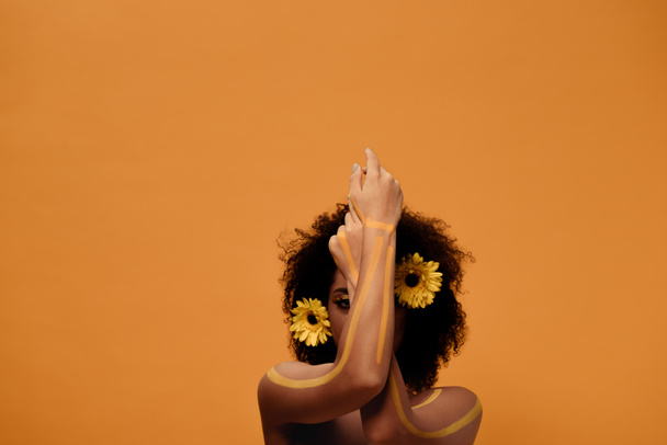 Молодая чувственная африканская американка с художественным макияжем и герберой в волосах, изолированных на оранжевом фоне
 - Фото, изображение