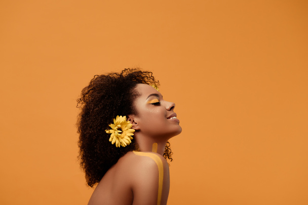 Stylish african american woman with artistic make-up and gerbera in hair isolated on orange background - Photo, Image