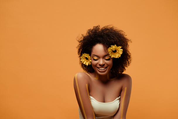 Jeune femme afro-américaine riante avec maquillage artistique et gerbera dans les cheveux isolés sur fond orange
 - Photo, image