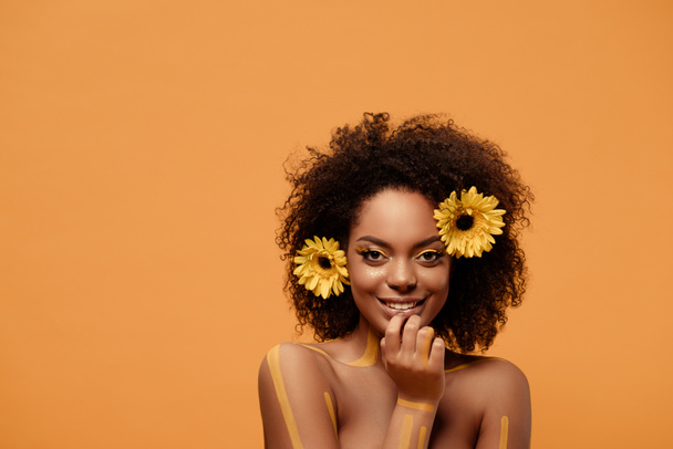 Jonge lachende african american vrouw met artistieke make-up en gerbera in haar geïsoleerd op een oranje achtergrond - Foto, afbeelding