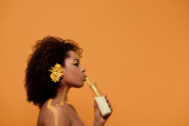 Young sensual african american woman with artistic make-up and gerbera in hair drinking milk from bottle isolated on orange background - Photo, Image