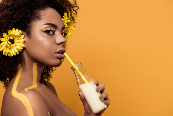 Привлекательная молодая африканская американка с художественным макияжем и герберами в волосах пьет молоко из бутылки, изолированной на оранжевом фоне
 - Фото, изображение