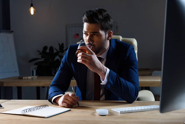 businessman drinking whiskey in dark office and looking away - Foto, afbeelding