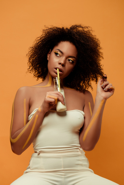 Молодая чувственная африканская женщина с художественным макияжем пьет молоко из бутылки, изолированной на оранжевом фоне
 - Фото, изображение