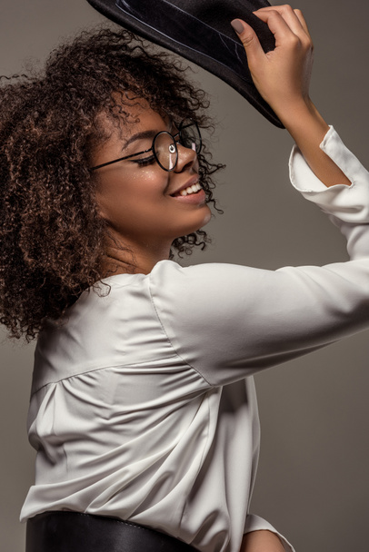 Attractive young american woman in white shirt wearing glasses pits black hat on isolated on grey background - Φωτογραφία, εικόνα
