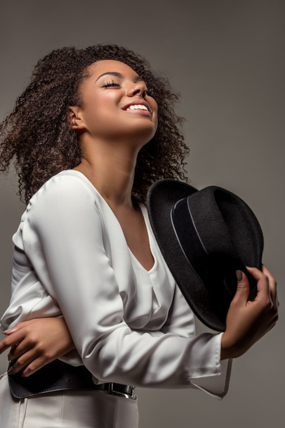 Gülümseyen ve siyah şapka gri arka plan üzerinde izole sallayarak Beyaz giysili şık Afrikalı-Amerikalı kadın - Fotoğraf, Görsel