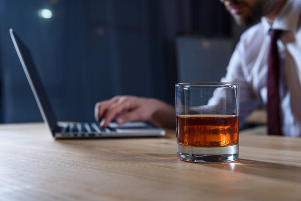 обрезанный образ бизнесмена, работающего за ноутбуком со стаканом виски на переднем плане
 - Фото, изображение