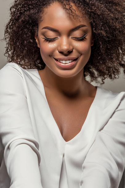 Κομψό αφρικανική αμερικανική γυναίκα σε άσπρο πουκάμισο χαμογελώντας και κοιτάζοντας προς τα κάτω απομονωμένες σε γκρι φόντο - Φωτογραφία, εικόνα