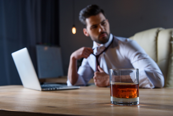 tired businessman loosen tie and sitting at table with glass of whiskey on foreground - Photo, Image