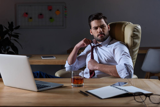 zmęczony biznesmen poluzować krawat i siedzi przy stole z szklanką whisky - Zdjęcie, obraz