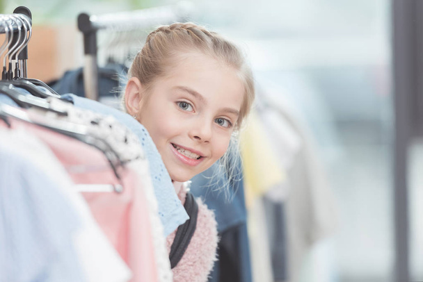 happy child looking out from row of clothes on hanger at shop    - Photo, Image