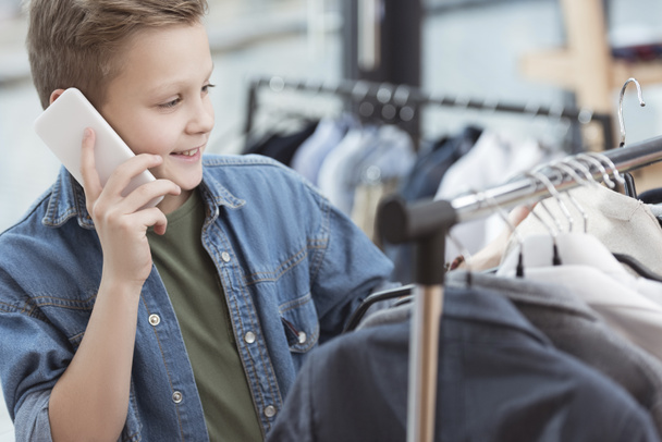 niño sonriente usando teléfono inteligente mientras sostiene la tela en la mano en la tienda
    - Foto, Imagen