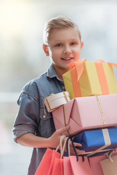 smiling boy holding boxes with colored paper bags in hands at shop interior    - Φωτογραφία, εικόνα