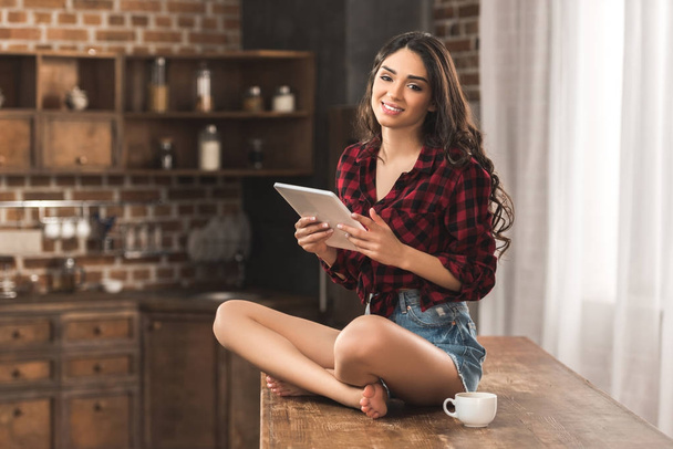 girl in checkered shirt and denim shorts smiling at camera while using digital tablet at home - Foto, afbeelding