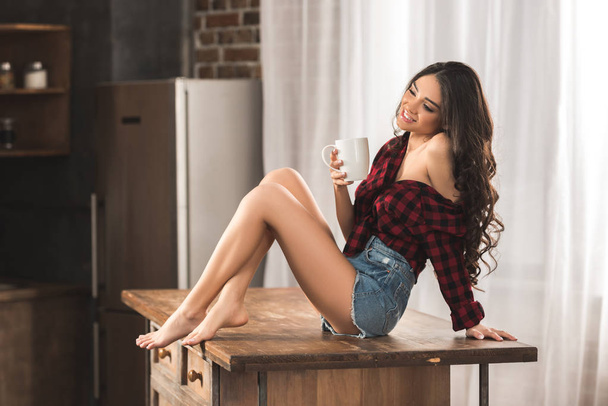 σέξι κορίτσι στο καρώ πουκάμισο και τζιν σορτς πίνοντας καφέ ενώ κάθεται στο τραπέζι στο σπίτι - Φωτογραφία, εικόνα