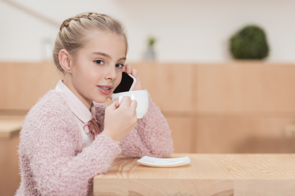 adorable niño sentado en la cafetería y hablando en el teléfono inteligente mientras bebe café en la cafetería
 - Foto, Imagen