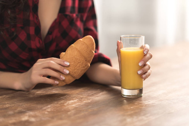 cropped shot of girl in checkered shirt holding croissant and glass of orange juice for breakfast - Foto, imagen