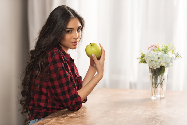mooi meisje in een geruit overhemd houden van groene appel en camera thuis te kijken - Foto, afbeelding