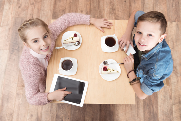 niños mirando la cámara mientras están sentados en la mesa con aparatos en la superficie en la cafetería
 - Foto, Imagen