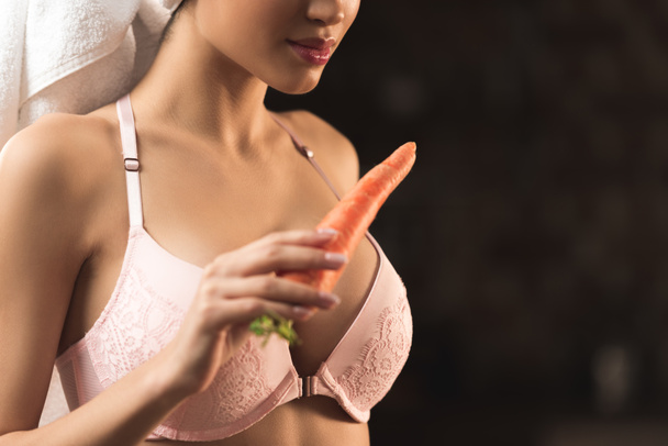 обрезанный снимок сексуальной молодой женщины в лифчике и полотенце, держащей морковь на черном
 - Фото, изображение