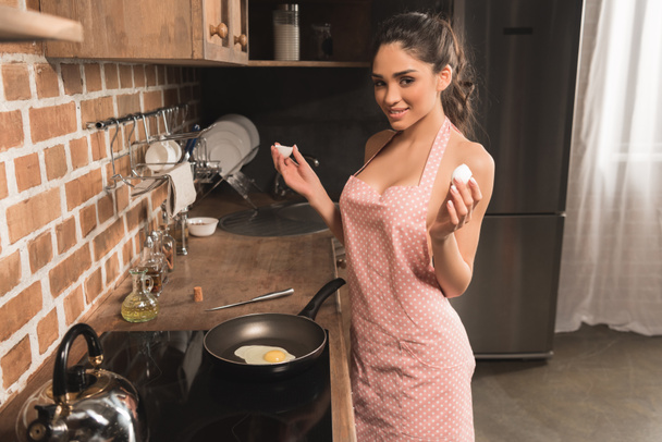 sexy joven en delantal sosteniendo cáscara de huevo y sonriendo a la cámara mientras se fríe el huevo en la cocina
 - Foto, Imagen