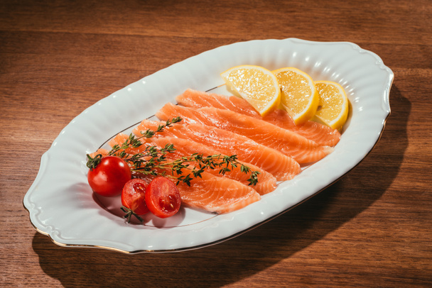 Salmon fish pieces with lemon, herbs and tomatoes on white plate on wooden table - Fotografie, Obrázek