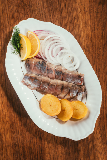Top view of herring fish pieces with lemon, onion and potato on white plate on wooden table - Fotografie, Obrázek