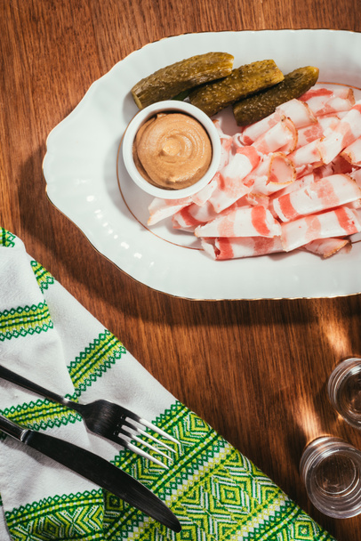 Top view of sliced bacon served with sauce and pickled cucumbers on plate served with drink on wooden table with napkin on wooden table - Photo, Image